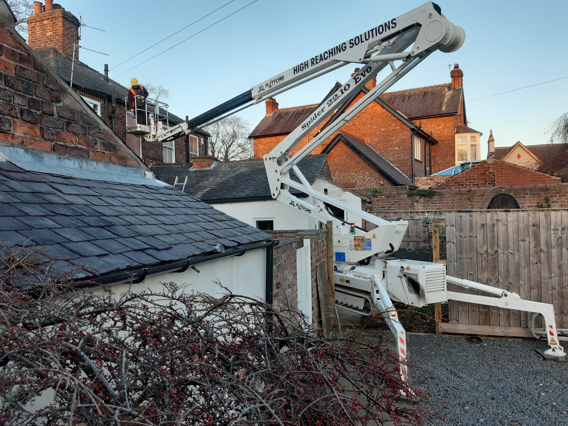 Tracked spider cherry picker set in middle of fence reaching over roof.